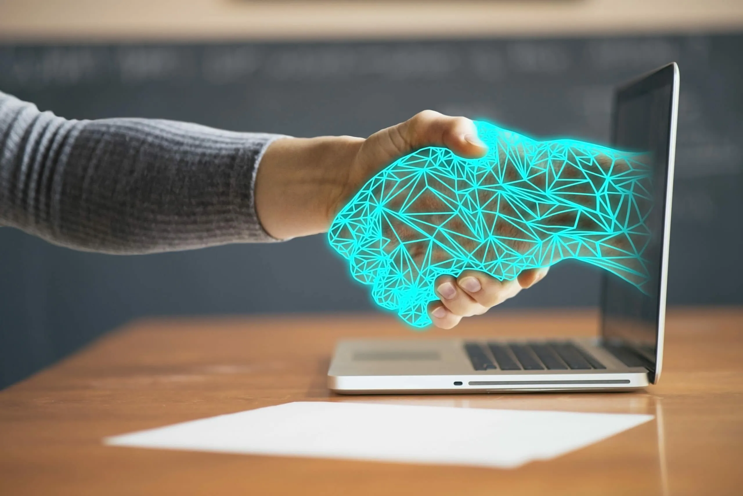 A hand shaking a glowing wireframe hand that reaches out of a laptop screen